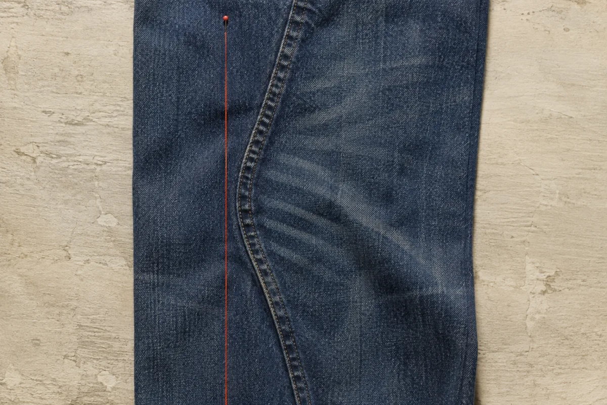 A washed denim leg shot straight on with its side seam curving away from the vertical, and a rust-red thread pinned down the line the seam was cut to follow
