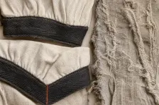 Two cream cuffs banded in dark cloth, one folded back to show the mitred join marked in rust-red, beside a third piece that is faded and frayed all over