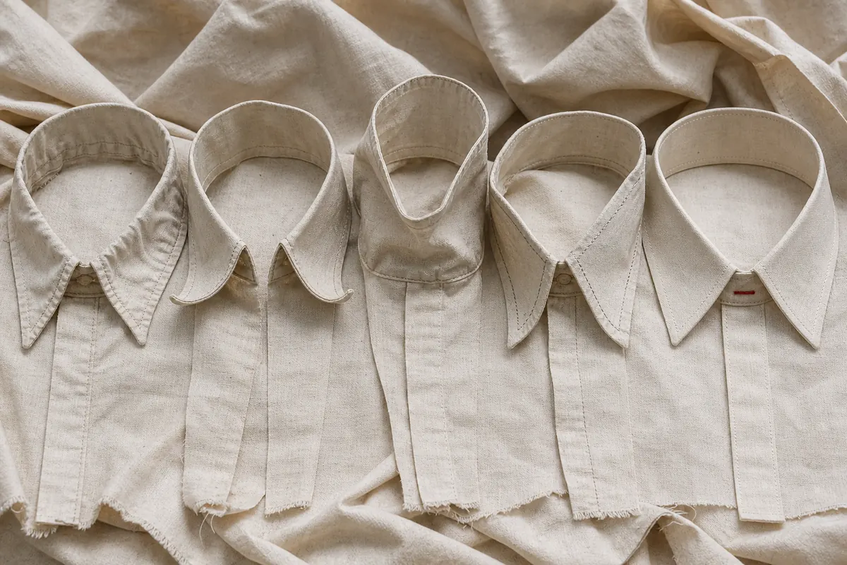 Five versions of the same calico shirt collar in a row: one puckered along the seam, one with the points curling up, one with the stand buckling, one with the topstitch wandering off the edge, and a fifth lying flat with a rust-red stitch at its centre